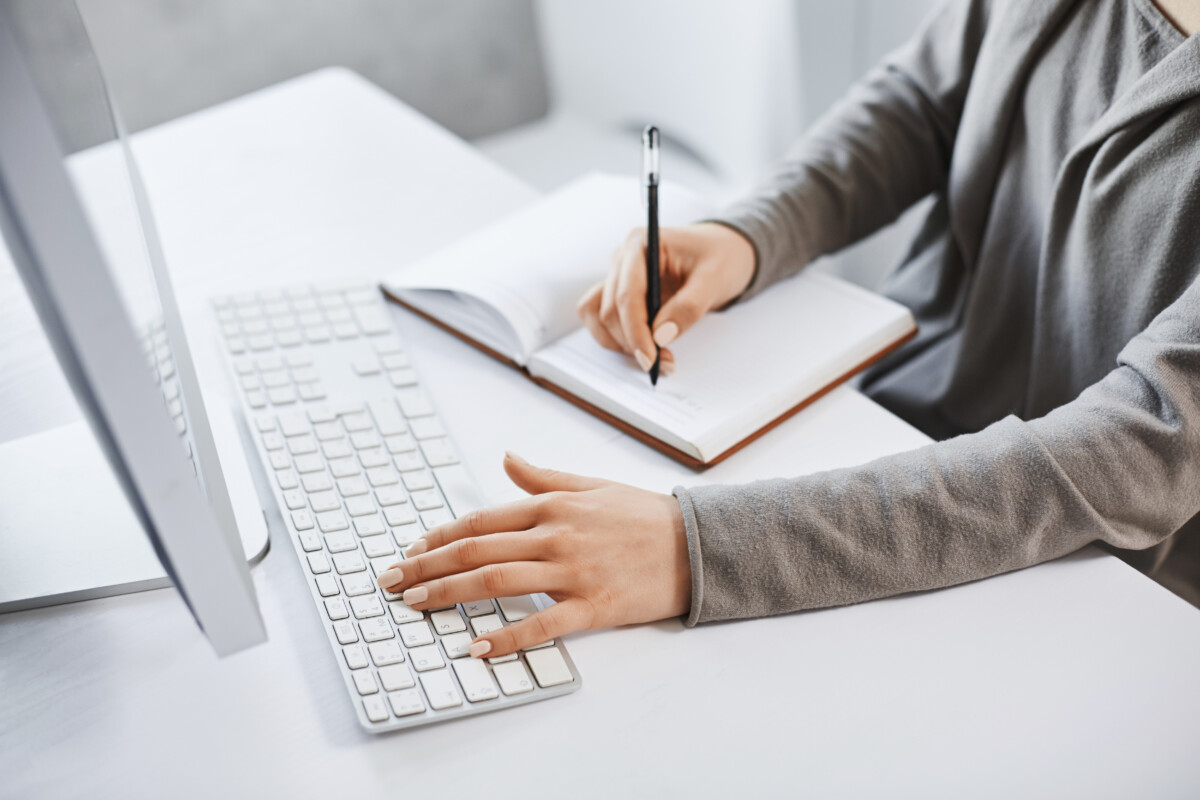 person working on computer while writing in notebook